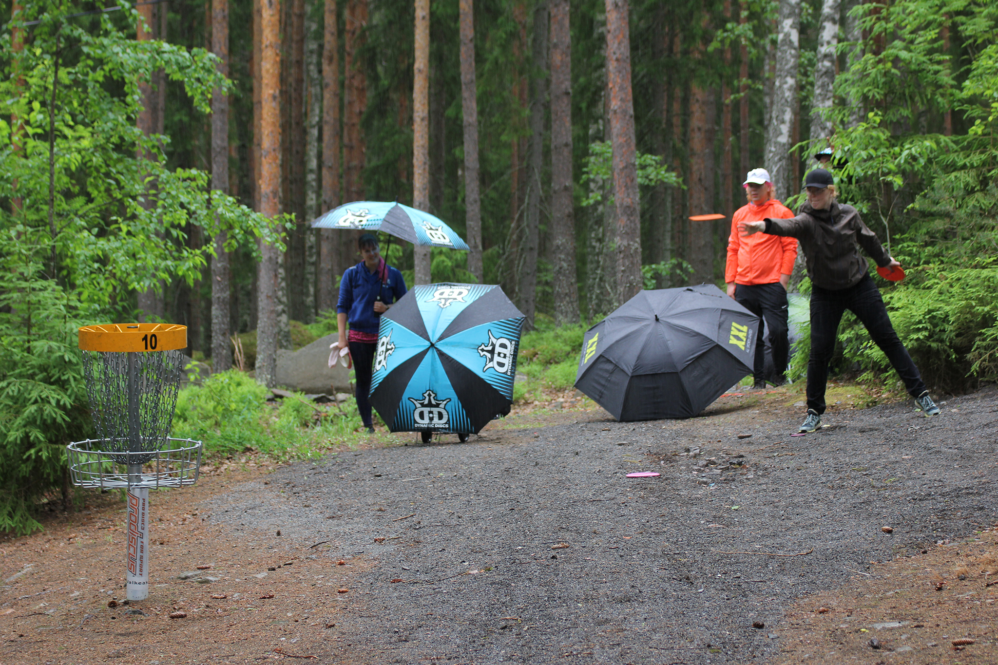 Kolme pelaajaa metsässä, sateenvarjot auki maassa, yhdellä kädessä. Yksi heittämässä.